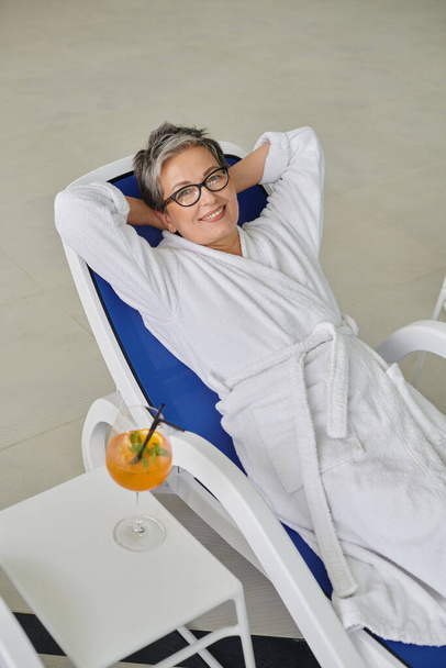 retraite concept, gelukkig volwassen vrouw in witte badjas rusten op ligstoel in de buurt van cocktail in spa centrum - Foto, afbeelding