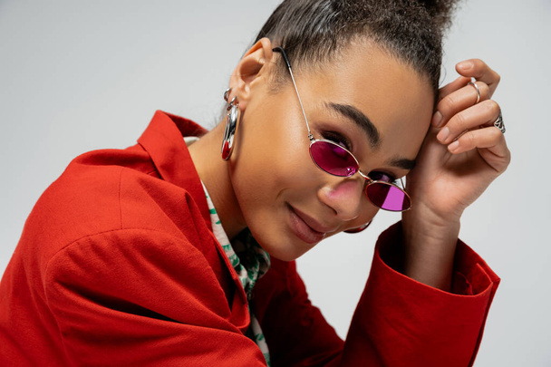 portrait of positive african american model in stylish attire and pink sunglasses on grey backdrop - Photo, Image