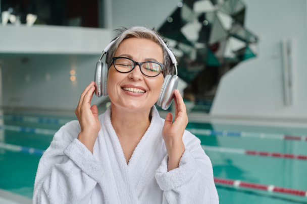 happy middle aged woman in glasses and robe listening music in wireless headphones in spa center - Photo, Image