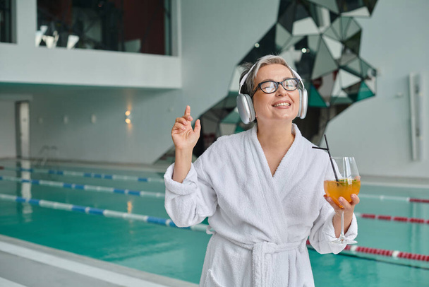 pleased middle aged woman listening music in wireless headphones and holding cocktail in spa center - Photo, Image