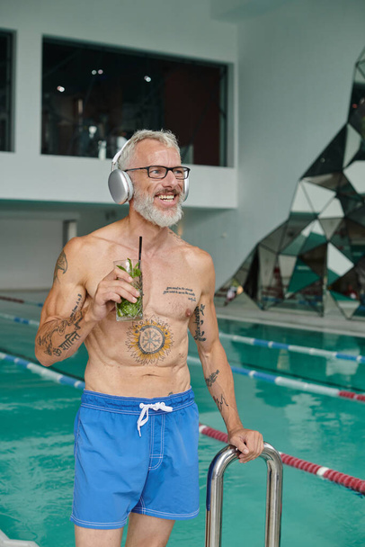 ретрит, татуировка, счастливый зрелый мужчина слушает музыку в наушниках и держит коктейль в спа - Фото, изображение