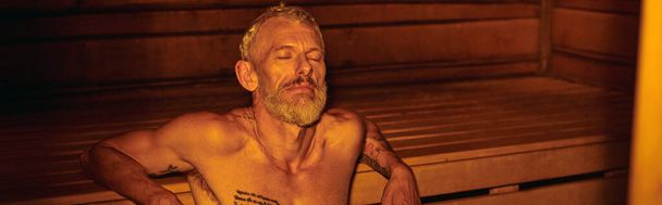 hombre tranquilo, relajado y sin camisa de mediana edad con tatuajes sentados en la sauna, el bienestar, la bandera - Foto, Imagen