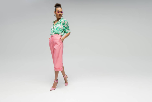 stylowa Afroamerykanka w różowych okularach przeciwsłonecznych i stylowym stroju pozowanie na szarym tle - Zdjęcie, obraz