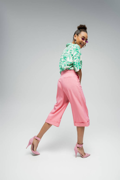 happy african american woman in pink sunglasses and stylish attire walking on grey backdrop - 写真・画像