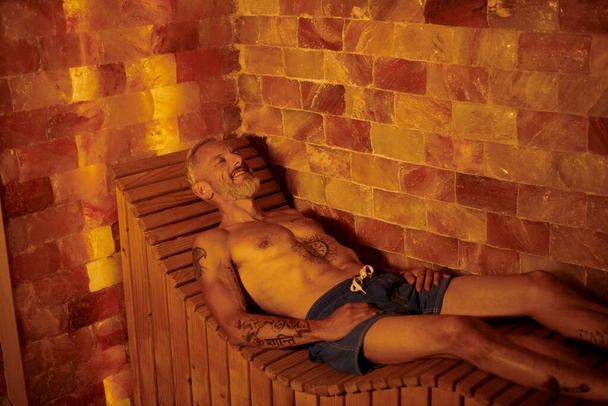 hombre de mediana edad alegre y sin camisa con tatuajes en la sauna, concepto de retiro de bienestar - Foto, Imagen