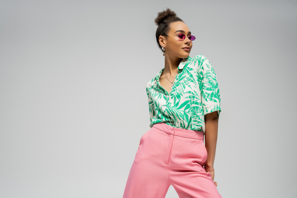 довольная африканская американка в розовых солнцезащитных очках и стильной одежде позирует на сером фоне - Фото, изображение