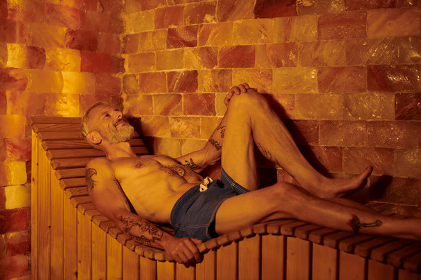 hombre de mediana edad relajado y sin camisa con tatuajes en la sauna, concepto de retiro de bienestar - Foto, Imagen