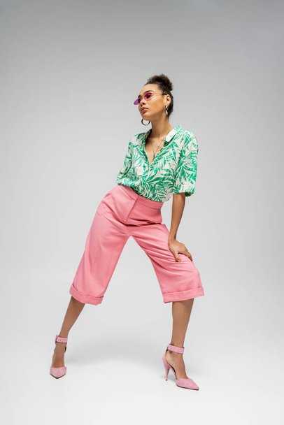 modelo de moda afroamericana de longitud completa con un atuendo elegante y tacones altos rosas sobre fondo gris - Foto, Imagen