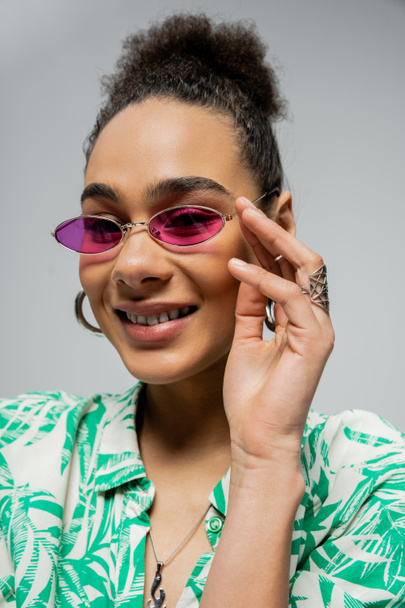 портрет позитивної афроамериканки в рожевих сонцезахисних окулярах і посмішка на сірому тлі - Фото, зображення