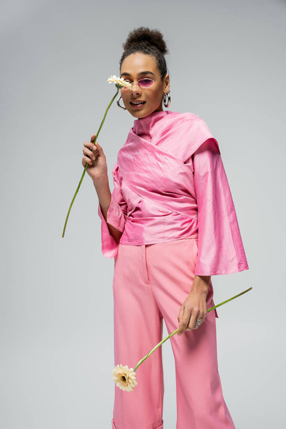 cheerful african american woman in pink attire and sunglasses posing with flowers on grey backdrop - Fotó, kép