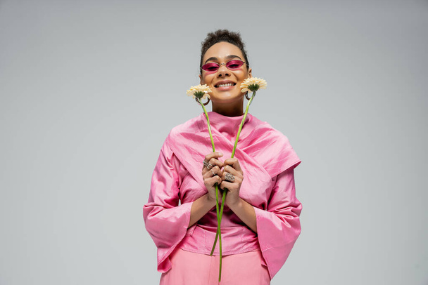 pleased african american woman in pink attire and sunglasses posing with flowers on grey backdrop - Φωτογραφία, εικόνα