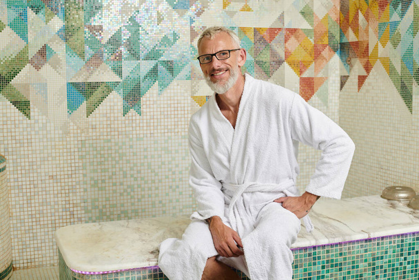 alegre hombre de mediana edad en bata blanca sentado en sauna, spa y concepto de bienestar, hammam de mármol - Foto, Imagen