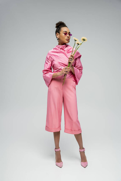 full length of african american woman in pink outfit and heels posing with flowers on grey backdrop - Zdjęcie, obraz