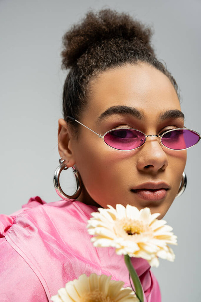 attractive african american fashion model in pink outfit and sunglasses posing with flowers on grey - Fotó, kép