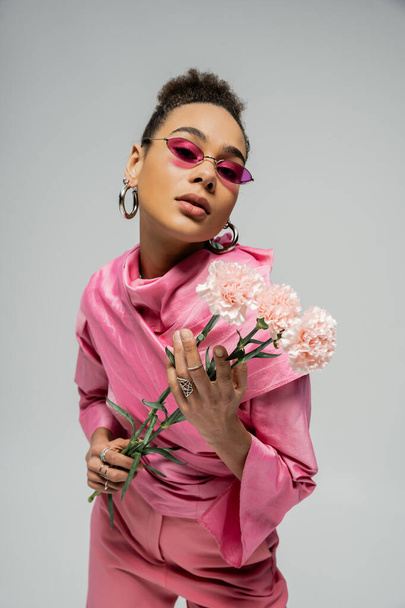 εκφραστικό αφρικανικό μοντέλο μόδας σε ροζ ενδυμασία και γυαλιά ηλίου με λουλούδια σε γκρι - Φωτογραφία, εικόνα