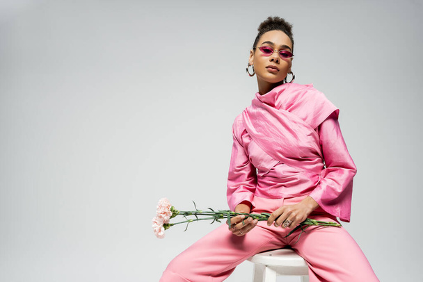 modelo afroamericano de moda en traje rosa y gafas de sol sosteniendo flores y sentado en la silla - Foto, Imagen
