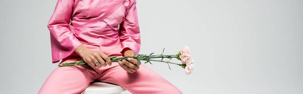 ořezaný módní model v růžovém oděvu držící květiny a sedící na židli, šedá pozadí banner - Fotografie, Obrázek