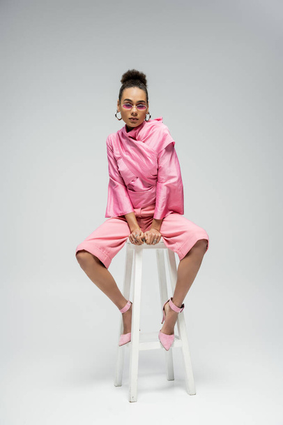 modelo afroamericano de moda en traje rosa y gafas de sol sentado en la silla y posando en gris - Foto, Imagen