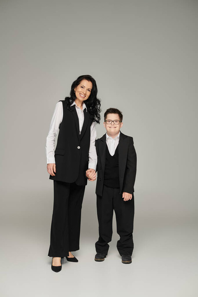 happy woman in formal wear holding hand of son with down syndrome in school uniform on grey - Foto, imagen