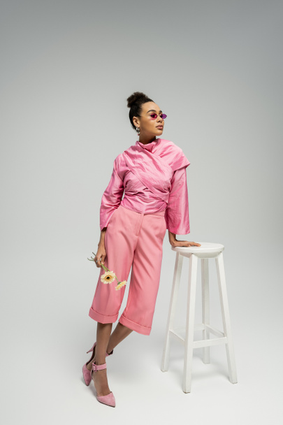 african american young woman in pink attire and sunglasses posing with flowers near stool on grey - Photo, Image