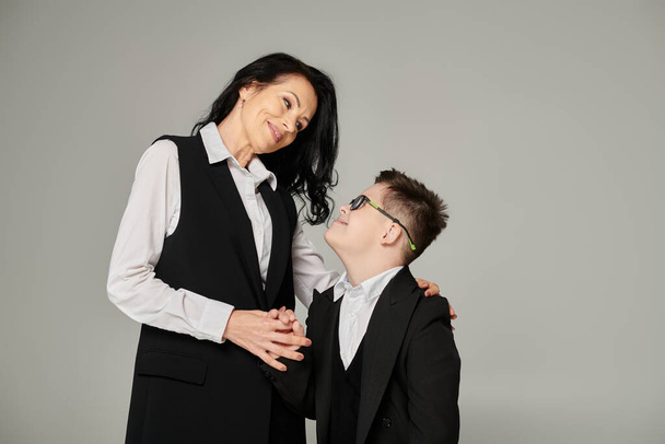 schoolboy with down syndrome and middle aged mother in formal wear smiling at each other on grey - Photo, Image