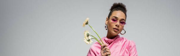 african american young woman in pink attire and sunglasses posing with flowers on grey, banner - Photo, Image