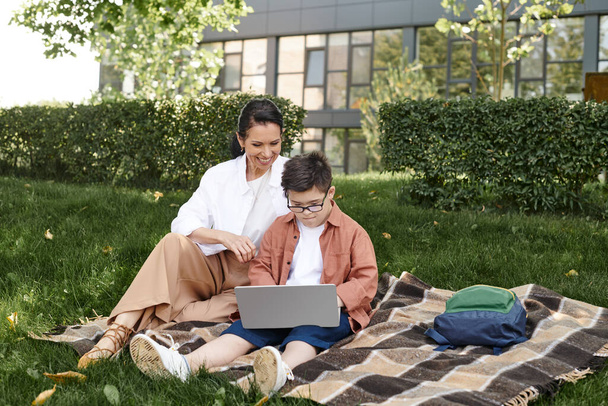 Junge mit Down-Syndrom, Brille, sitzt mit Laptop im Park neben glücklicher Mutter, E-Learning - Foto, Bild