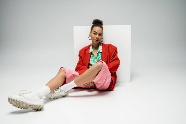 pretty african american woman in fashionable and vibrant outfit sitting near cube on grey backdrop - Foto, Imagem