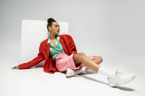 pretty african american woman in modern and vibrant clothes sitting near cube on grey backdrop - Photo, Image