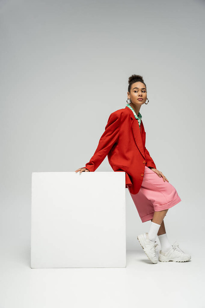 attractive african american woman in vibrant outfit leaning on cube on grey backdrop, look away - Foto, imagen