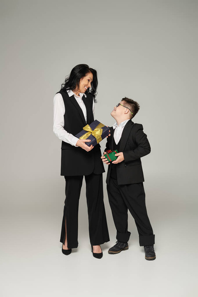 бізнес-леді і щасливий син з синдромом Дауна в шкільній формі тримає подарункові коробки на сірому - Фото, зображення