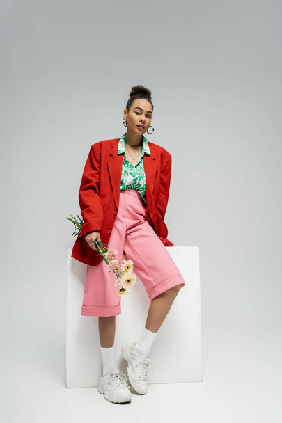 african american woman in stylish red blazer holding flowers and leaning on cube on grey backdrop - 写真・画像