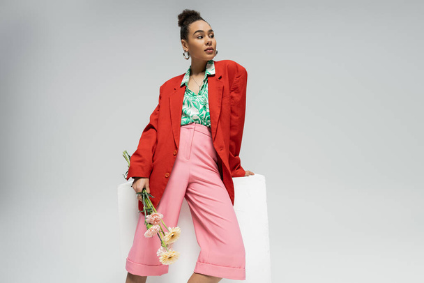 pretty african american woman in red blazer holding flowers and leaning on cube on grey backdrop - Photo, Image