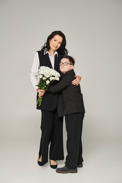 Ευτυχισμένη γυναίκα και αγόρι με σύνδρομο Down κρατώντας λουλούδια και αγκαλιάζοντας μια γκρίζα, μοναδική οικογένεια - Φωτογραφία, εικόνα