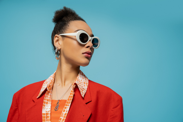 Porträt einer afrikanisch-amerikanischen Frau mit Reifrohren, Sonnenbrille und lebendigem Outfit, die auf blauem Grund posiert - Foto, Bild