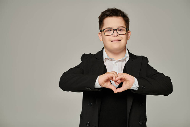garçon heureux avec le syndrome du duvet en uniforme scolaire et lunettes montrant signe d'amour avec les mains sur gris - Photo, image