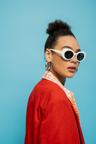 eye-catching african american model in vibrant outfit and sunglasses on blue background, portrait - Photo, Image