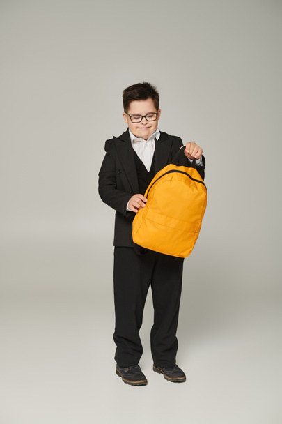 happy schoolboy with down syndrome, in black uniform and eyeglasses holding yellow backpack on grey - Photo, Image