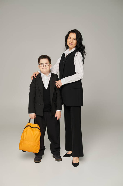 heureux écolier avec le syndrome du duvet, avec sac à dos jaune près et mère en tenue formelle sur gris - Photo, image