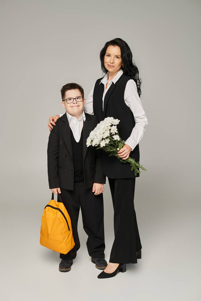 woman with flowers and schoolboy with down syndrome holding yellow backpack on grey, special family - Photo, Image