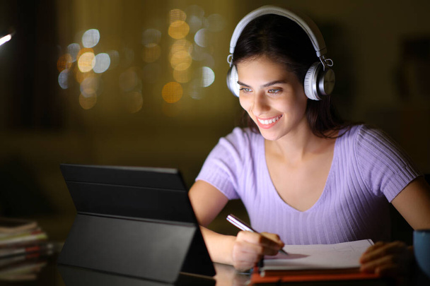 Happy student using tablet and headphone and taking notes in the night at home - 写真・画像