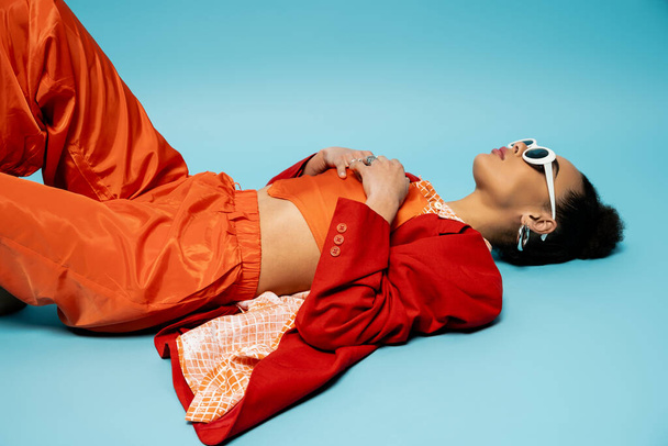 attraktives afrikanisch-amerikanisches Model in stilvollem, lebendigem Outfit auf blauem Hintergrund liegend, entspannte Pose - Foto, Bild