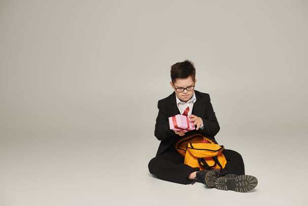 школьник с синдромом Дауна держит подарочную коробку и сидит рядом с желтым рюкзаком на сером, полной длины - Фото, изображение