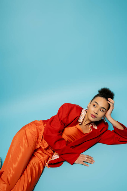 pretty african american model in silver accessories and vibrant outfit lying on blue backdrop - 写真・画像