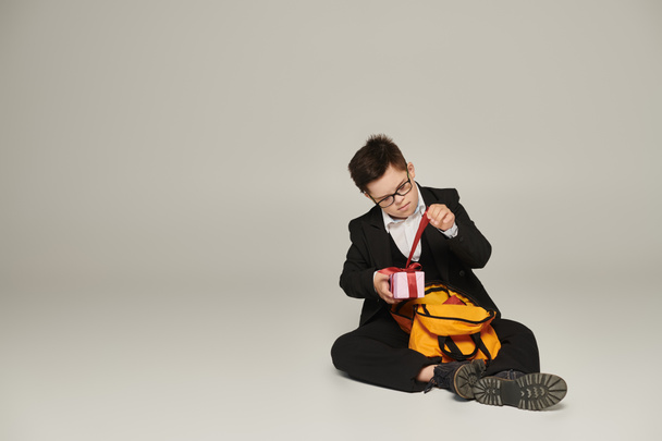 colegial con síndrome de Down, en uniforme negro sentado cerca de la mochila y abriendo la caja de regalo en gris - Foto, Imagen
