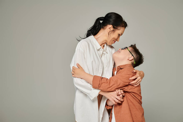 niño con síndrome de Down y mujer de mediana edad en ropa de moda abrazando en gris, familia única - Foto, Imagen