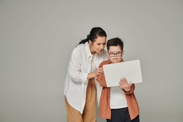Женщина средних лет объясняет онлайн урок улыбающемуся сыну с синдромом Дауна рядом с ноутбуком на сером - Фото, изображение