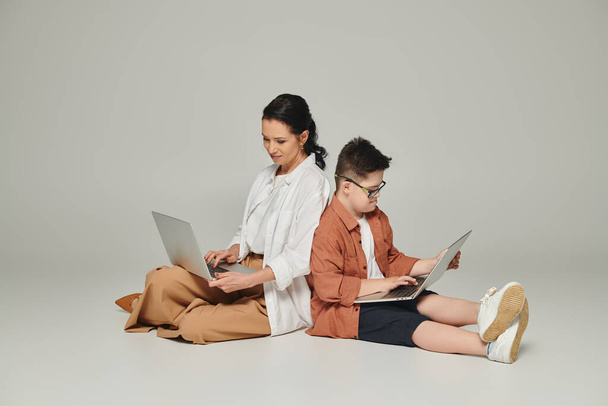 kobieta w średnim wieku i dziecko z zespołem Downa siedzi plecami do siebie z laptopami na szarym - Zdjęcie, obraz