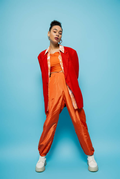 gorgeous fashion model in bright orange clothing looking at camera and posing on blue background - Foto, Imagem