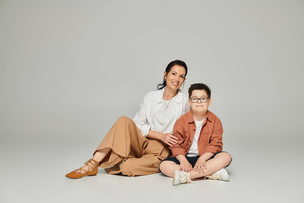 жінка середнього віку і хлопчик з синдромом Дауна сидячи і дивлячись на камеру на сірій, особливій сім'ї - Фото, зображення